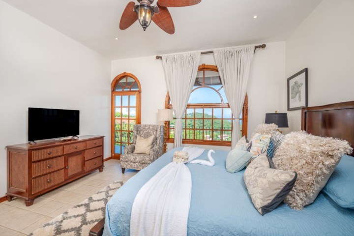 Stylish bedroom with blue bedding, ornate pillows, TV, and large windows with curtains, overlooking greenery.