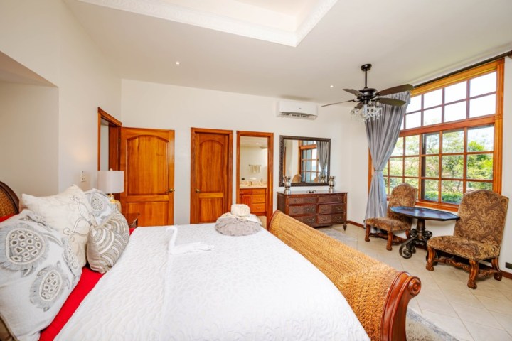 Cozy bedroom with large bed, wooden doors, dresser, and sitting area by a window with garden view.
