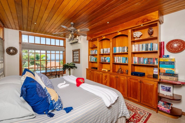 Cozy bedroom with wooden shelves, books, and board games, large window with sea view, ceiling fan overhead.