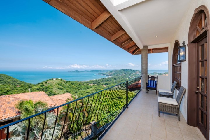 Balcony with seating, ocean and lush green hills in the distance on a clear day.