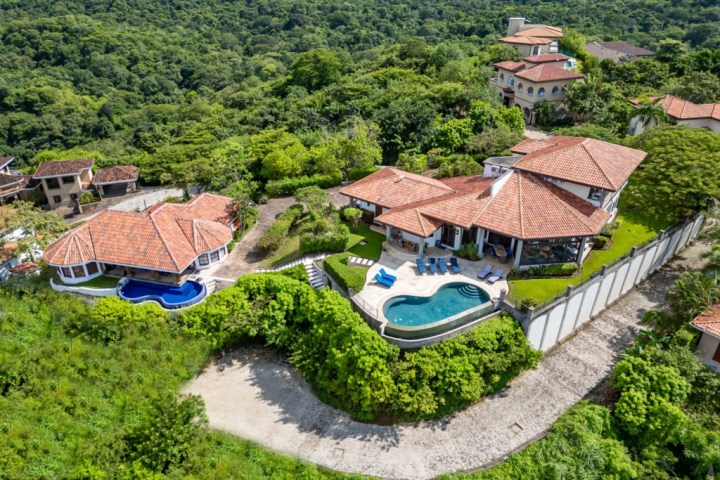 Aerial view of a large house with a pool surrounded by lush greenery and other homes.