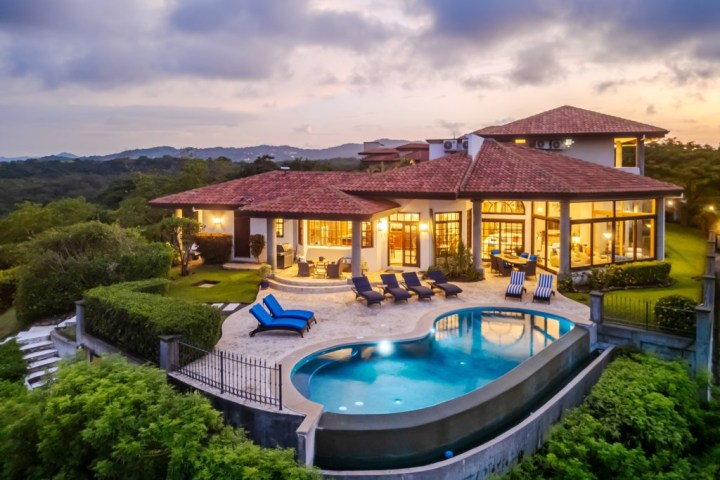 Luxury house with red roof, curved pool, lounge chairs, and lush greenery at sunset.