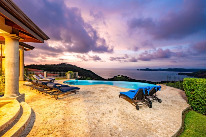 Infinity pool and loungers overlook ocean and hills at sunset.