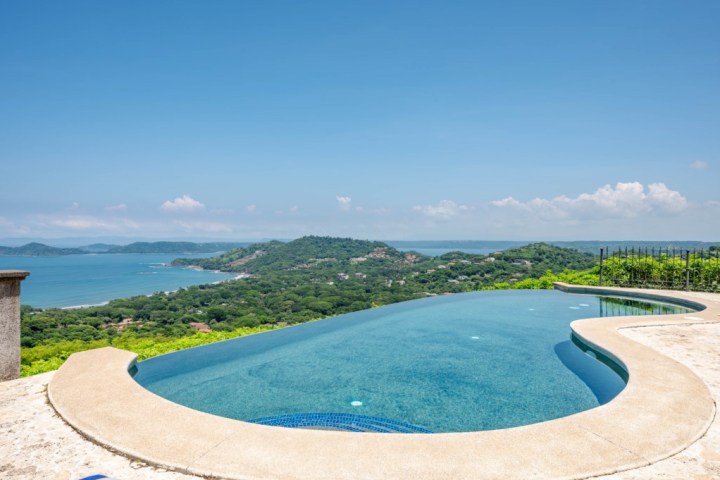 Infinity pool with ocean and lush hillside views under a clear blue sky.