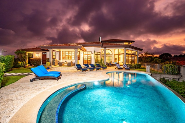 Luxury house with large windows, pool, and lounge chairs under a dramatic evening sky.