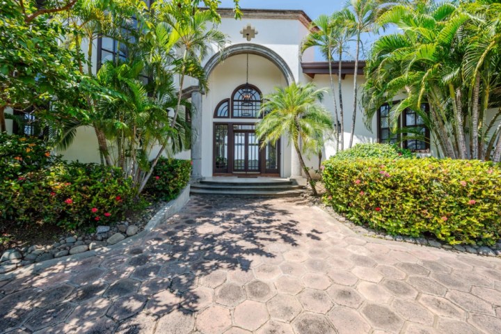 Large house entrance with arched doorway, surrounded by palm trees and shrubs.