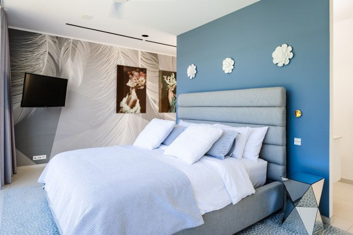 Modern bedroom with blue accent wall, decorative pillows, wall-mounted TV, and abstract wall art.
