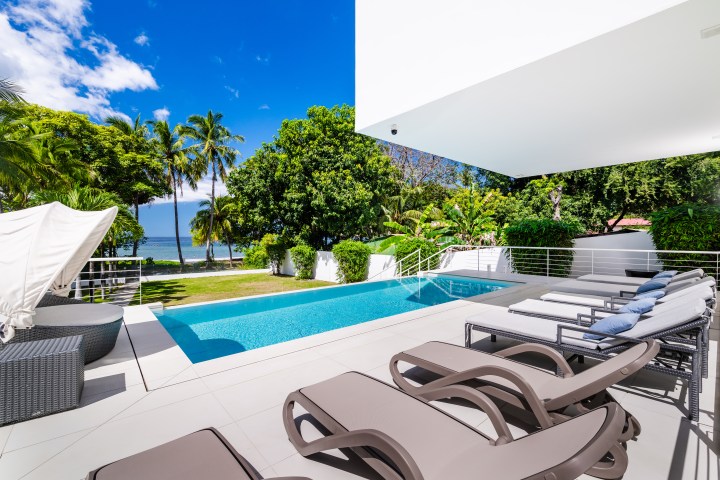 Modern outdoor pool area with lounge chairs, palm trees, ocean view, and clear blue sky.