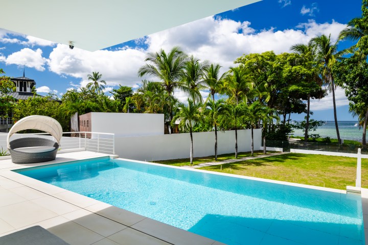 Modern poolside area with lounge chair, palm trees, ocean view, and partly cloudy sky.