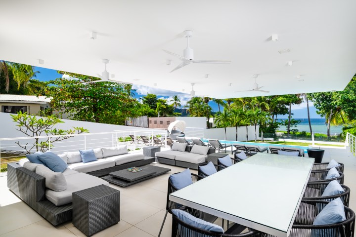 Modern outdoor patio with seating, glass table, ceiling fans, and view of pool and ocean.