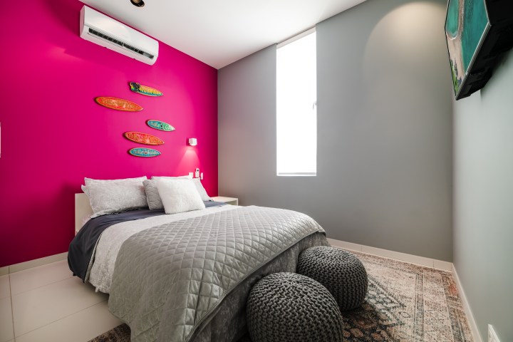 Bedroom with pink accent wall, surfboard decor, gray bedding, and white window shade.