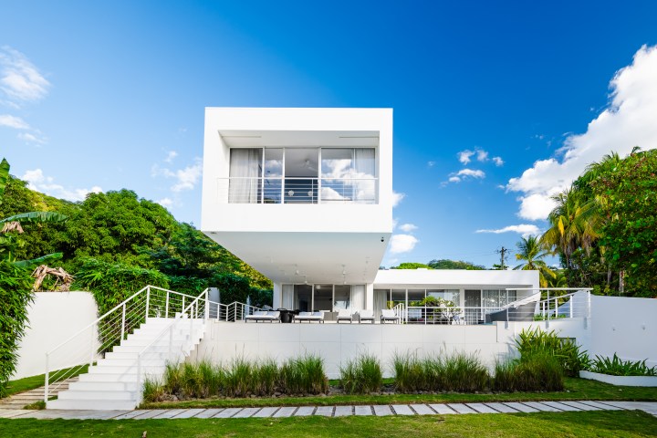 Modern white house with large windows, greenery around, and a blue sky background.