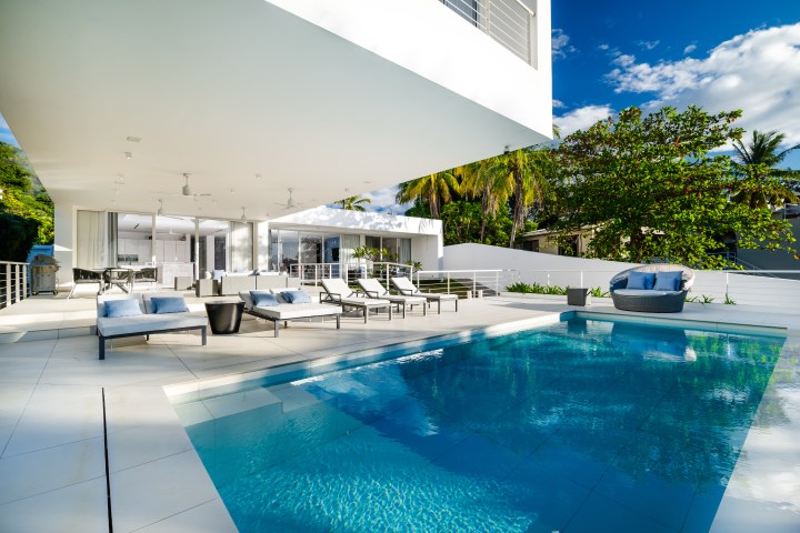 Modern outdoor patio with pool, lounge chairs, and a covered seating area under a clear blue sky.