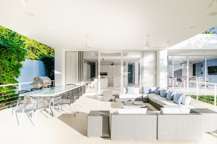 Modern patio with wicker furniture, dining table, and grill, adjacent to open kitchen, lush greenery around.