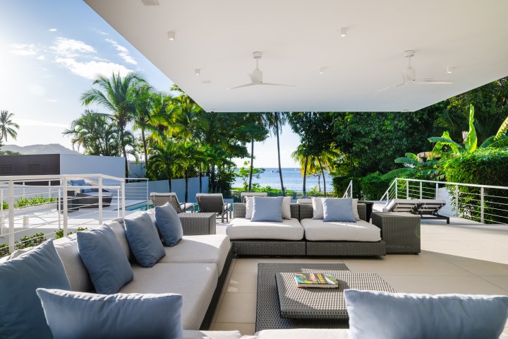 Beachfront patio with gray sofas, blue cushions, and ocean view surrounded by palm trees.