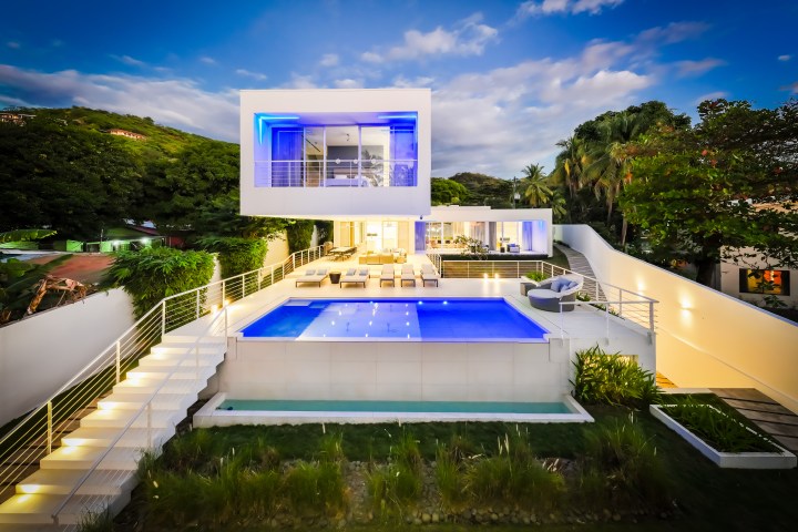Modern house with illuminated pool and stairs, surrounded by trees at dusk.