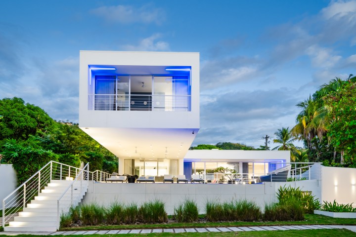 Modern white house with blue lights, surrounded by trees and a landscaped garden.