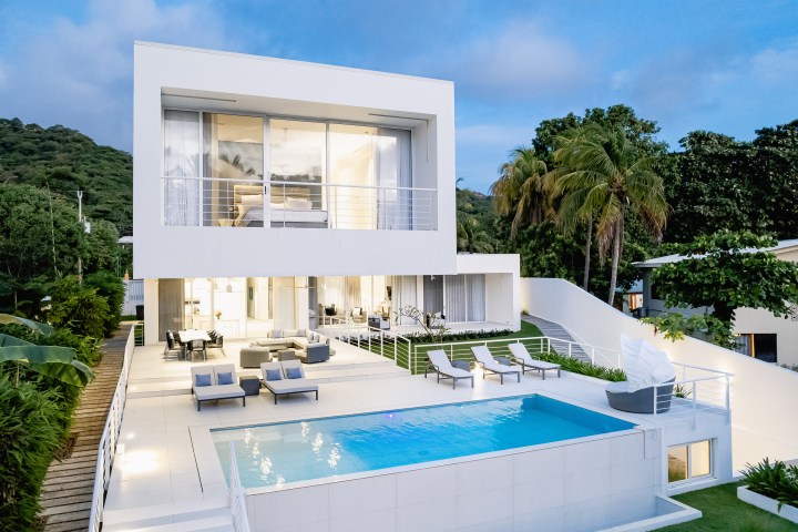 Modern white house with large windows, patio furniture, and swimming pool surrounded by trees.