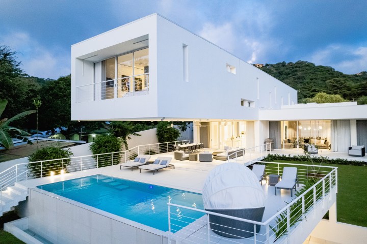 Modern white house with large windows, balcony, pool, and mountain backdrop.