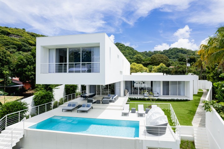 Modern white villa with pool, terrace, and garden, surrounded by lush greenery and a blue sky.