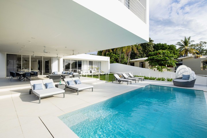 Modern patio with lounge chairs, dining table, and a pool, surrounded by greenery and a white building.