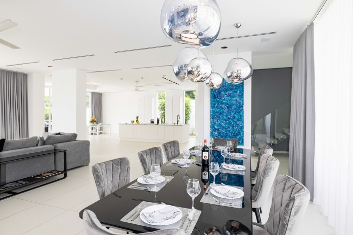 Modern dining room with mirrored table, grey chairs, and pendant lights, open to kitchen and living area.