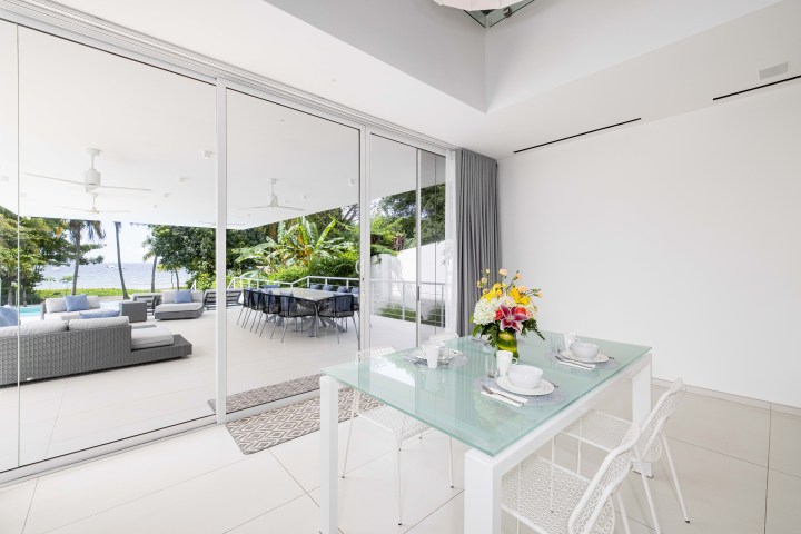Modern dining area with glass table, flowers, and large windows overlooking patio with sofa and trees.