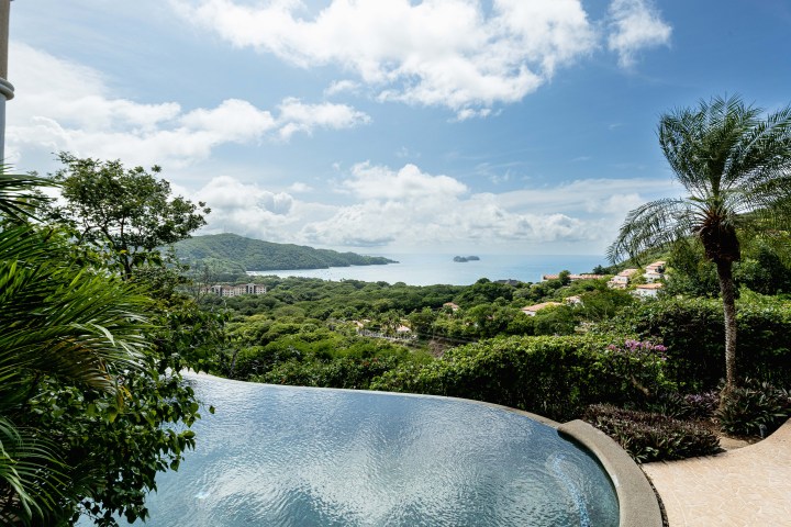 Infinity pool overlooking lush hills and ocean under a cloudy blue sky.