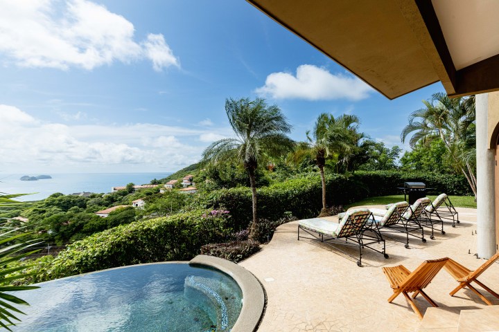 Patio with pool, lounge chairs, and ocean view under a blue sky.
