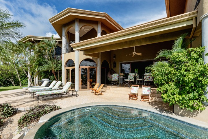 Luxury villa patio with pool, lounge chairs, and tropical plants under a blue sky.