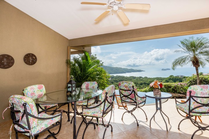 Outdoor patio with floral chairs, glass table, ceiling fan, and ocean view with lush greenery.