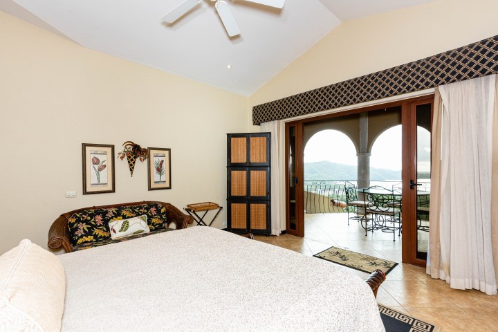 Bedroom with bed, sofa, framed art, and open doors to balcony with mountain view.