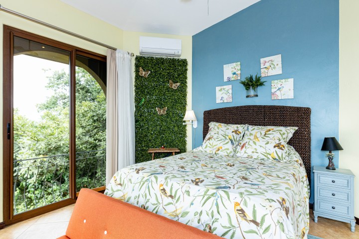 Bedroom with floral bedding, blue wall, leafy decor, artwork, and large window with trees outside.
