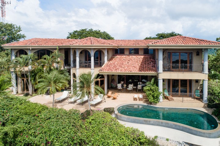 Luxury villa with red-tiled roof, pool, and palm trees in front of balconies.