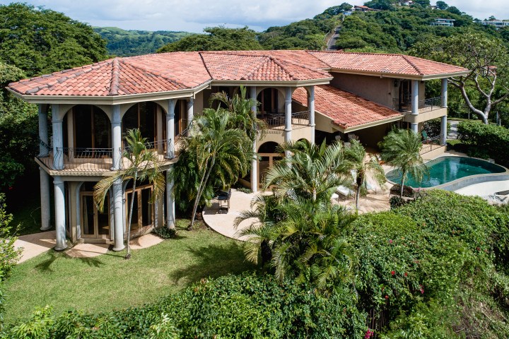 A large villa with a red-tiled roof, columns, pool, and lush greenery.
