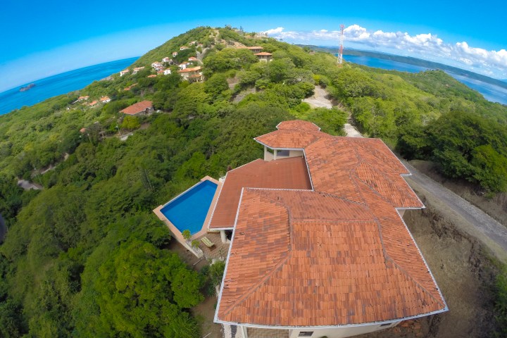Aerial view of a coastal house with red roof and blue pool surrounded by lush greenery.