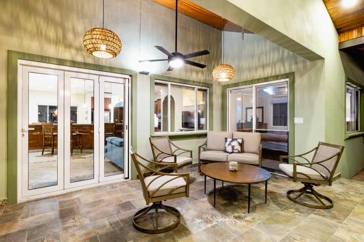 Outdoor patio space with chairs, table, overhead fan, and lights, next to sliding glass doors and windows.