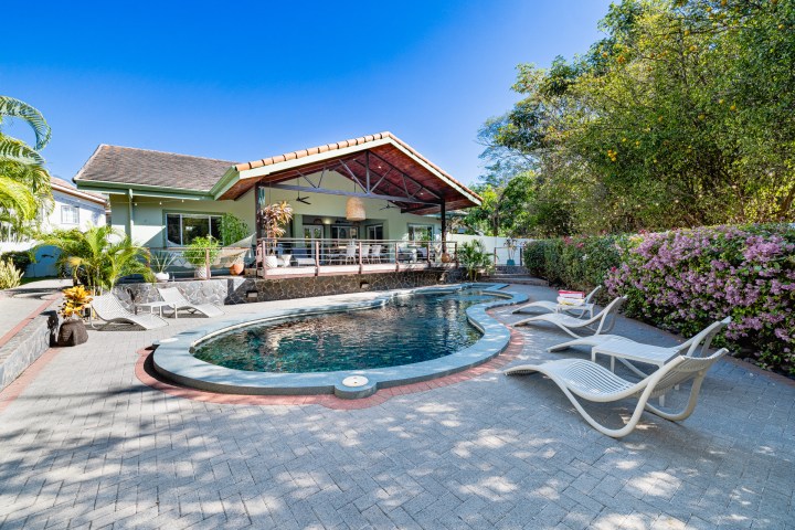 House with patio and pool, surrounded by trees and plants, under a clear blue sky.