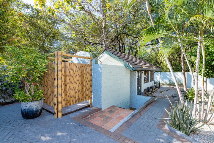 Small building with bamboo shower and tropical plants in a sunny garden setting.