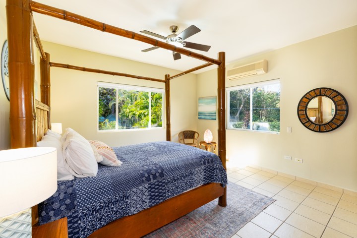 Bright bedroom with bamboo canopy bed, ceiling fan, and large windows overlooking greenery.