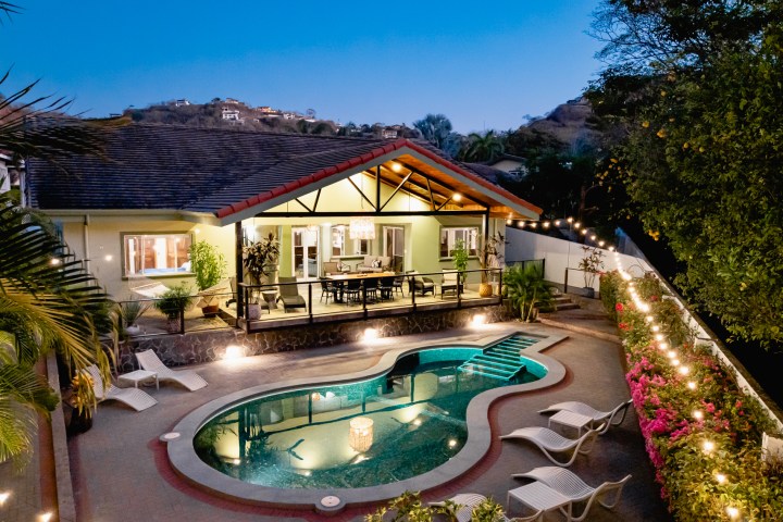 Illuminated backyard with pool, patio, and house at dusk, string lights overhead.