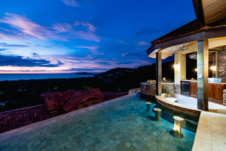 Infinity pool with ocean view at sunset, near a luxurious house with lit pillars.