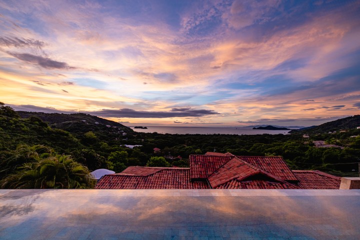Scenic sunset view over rooftops with trees and water in the background.