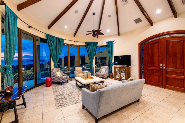 Spacious living room with blue curtains, tiled floor, and arched wooden ceiling beams.