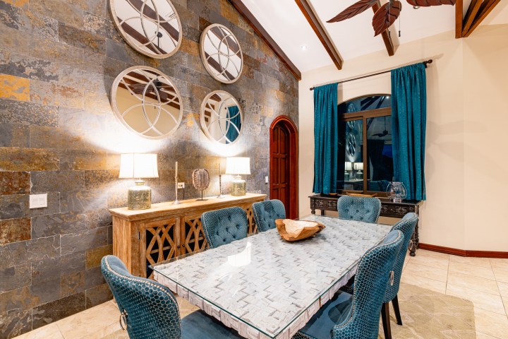 Elegant dining room with blue chairs, glass table, round mirrors, and teal curtains.
