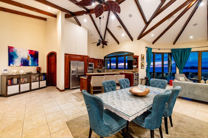 Spacious modern living room with dining area, open kitchen, tall ceiling, and sunset view.