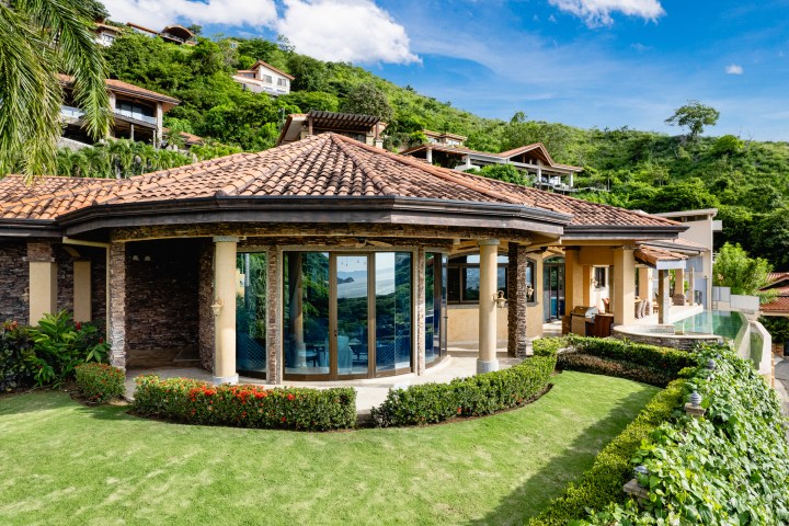 Villa with tiled roof, large windows, garden, and hillside background on a sunny day.
