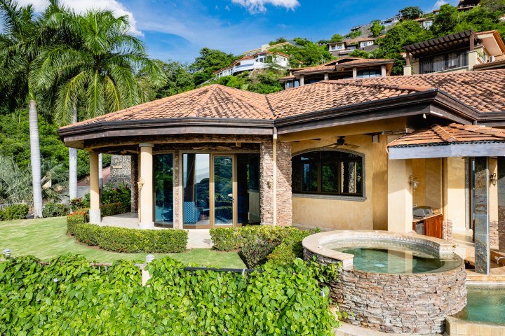 Tropical villa with pool, stone wall, and lush greenery under a blue sky.