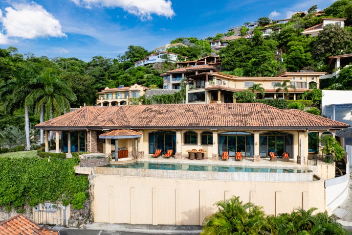Hillside villa with a tiled roof, pool, surrounded by lush greenery.