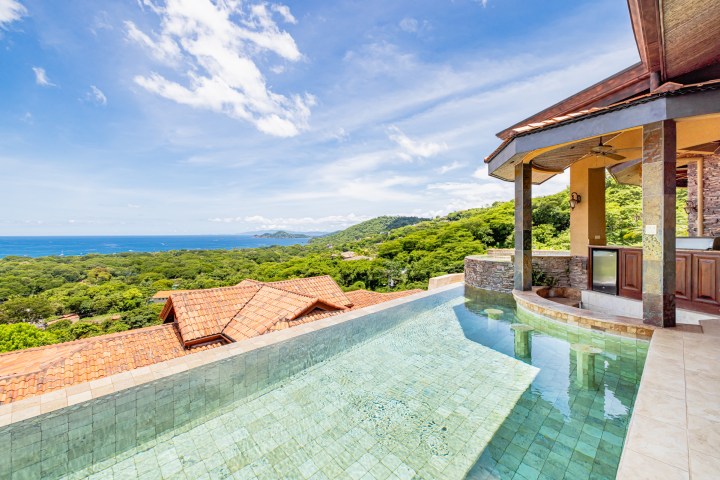 Infinity pool with ocean and green landscape view under a clear blue sky.
